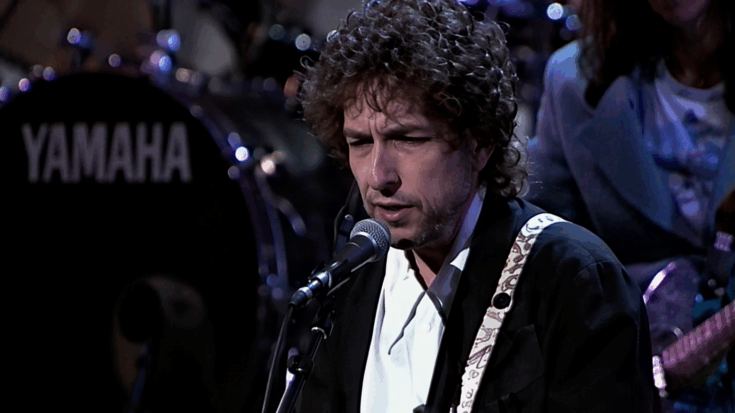 Bob Dylan sings into a microphone onstage, wearing a dark jacket with a focused, serious expression.
