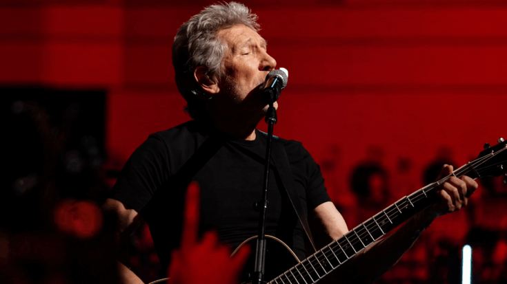 Roger Waters sings into a microphone while playing guitar, set against a bold red-lit stage backdrop.