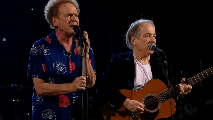 Simon & Garfunkel perform together onstage, with one singing into a microphone while the other plays acoustic guitar.