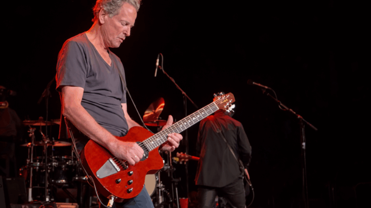 Lindsey Buckingham plays a bright red guitar onstage under warm red lights with calm focus.