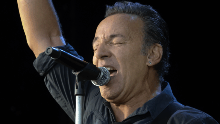 Bruce Springsteen sings passionately into the microphone with one arm raised high onstage.