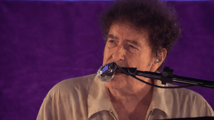 Bob Dylan sings at the piano in a close-up performance shot under soft purple light.