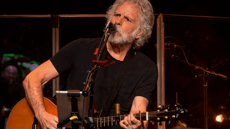 Bob Weir sings into the microphone while playing guitar onstage with a focused expression.