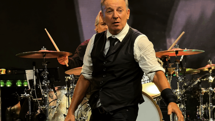 Bruce Springsteen strides across the stage in a sharp black vest and tie while the drummer plays behind him.