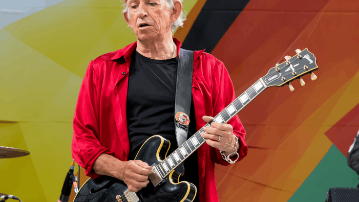 Keith Richards plays his black guitar onstage while wearing a bright red shirt and matching beanie.