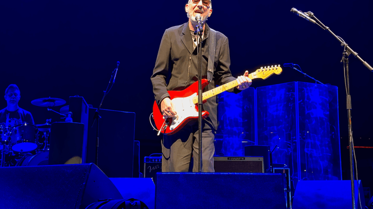 Pete Townshend performs onstage with a red guitar under deep blue concert lights.