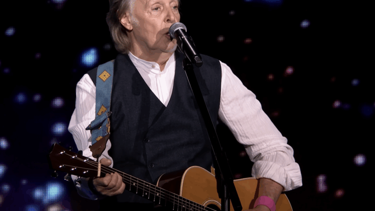 Paul McCartney sings into the microphone while playing an acoustic guitar against a glowing dark stage backdrop.