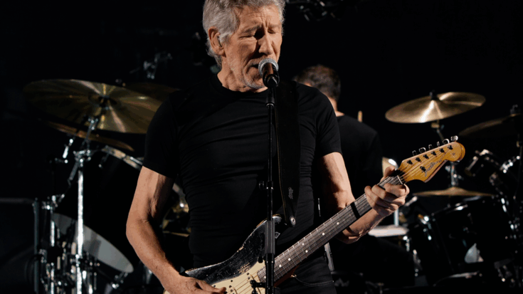 Roger Waters sings into the microphone while playing guitar under dark concert lights.