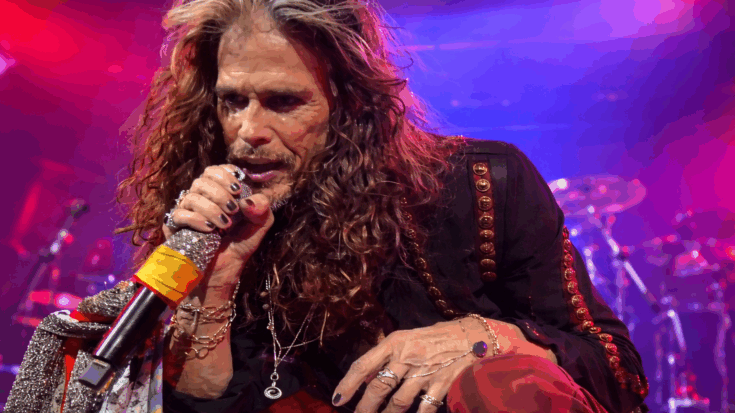 Steven Tyler kneels onstage, gripping a sparkling microphone under vivid concert lights.