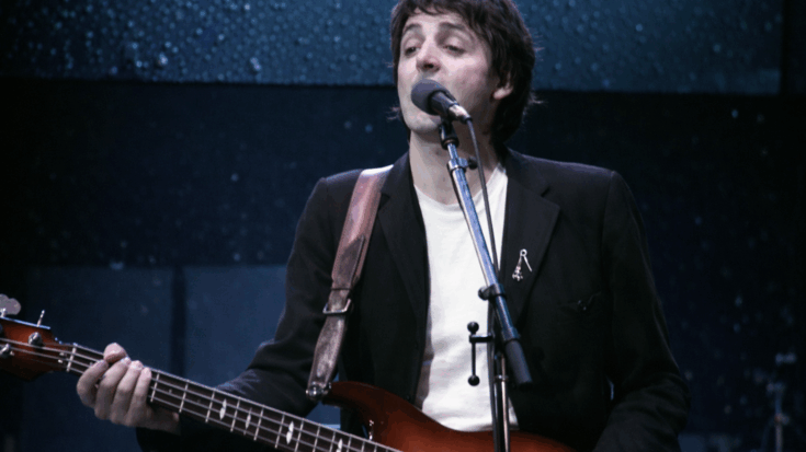 Paul McCartney sings into a microphone while playing his sunburst bass guitar onstage.
