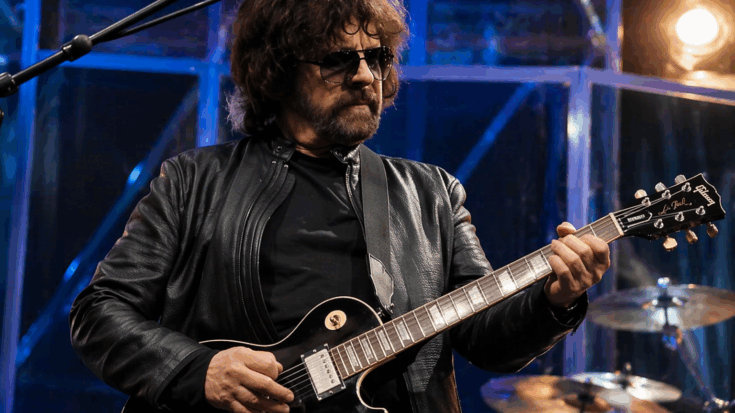 Jeff Lynne stands onstage in dark sunglasses, playing a black electric guitar under bright concert lights.