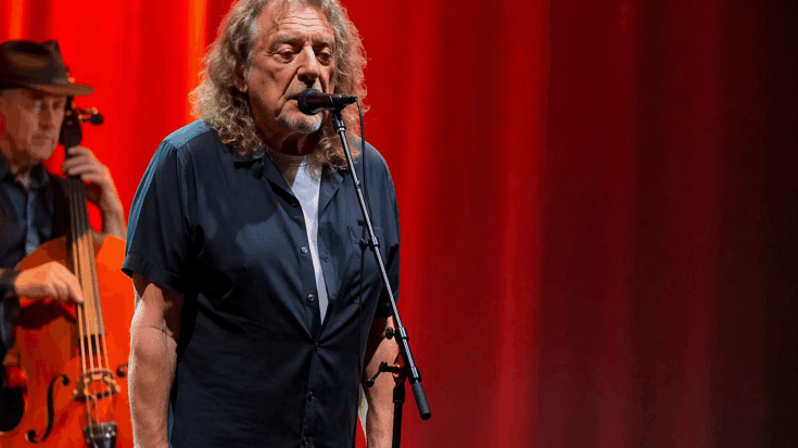 Robert Plant stands at a microphone under glowing red stage lights during a live performance.