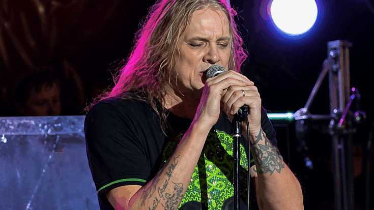 Sebastian Bach sings intensely into the microphone under colorful stage lights, wearing a skull shirt and showing tattooed arms.