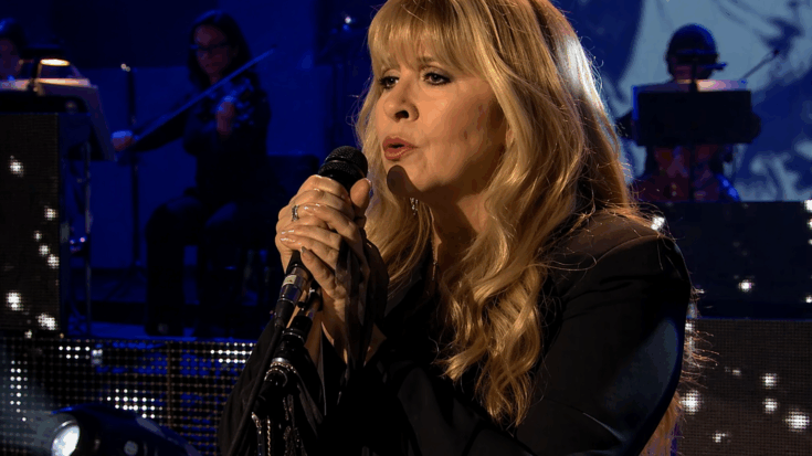 Stevie Nicks sings softly into the microphone under blue stage lights, surrounded by an elegant concert setting.