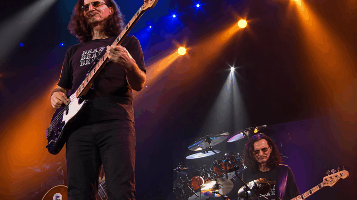 Geddy Lee plays bass onstage under bright orange and blue lights, surrounded by a powerful live concert setup.