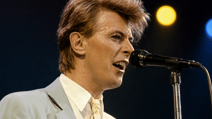 David Bowie sings into the microphone in a sharp suit, glowing under warm stage lights during a classic live performance.