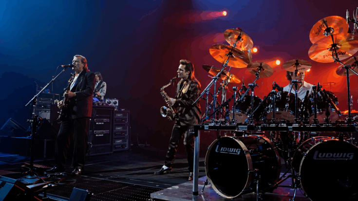 Pink Floyd performs on a dimly lit stage with rich red tones, surrounded by drums, instruments, and a powerful concert atmosphere.