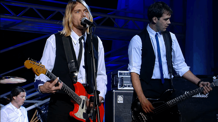 Kurt Cobain sings into the microphone while playing guitar onstage, surrounded by cool blue concert lighting.