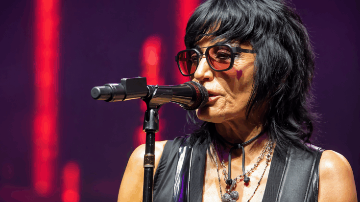 Joan Jett sings into the microphone under vivid red stage lights, wearing dark shades and her signature rock style.