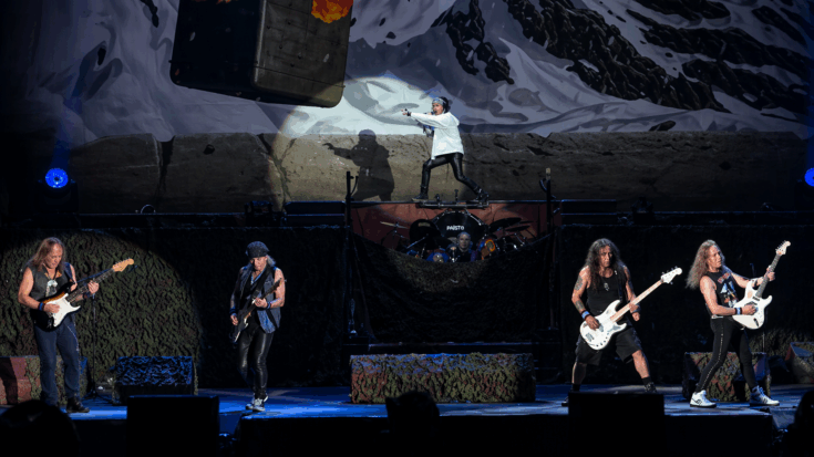 Iron Maiden performs on a massive stage with dramatic lighting, a mountain backdrop, and the band spread across the scene in full concert mode.