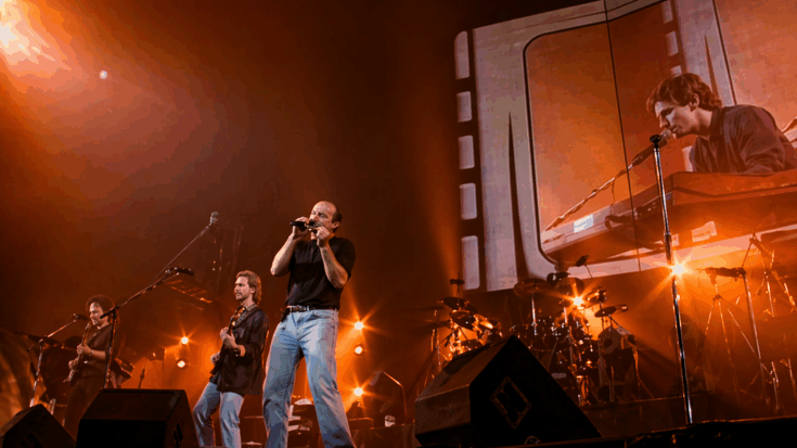 The Genesis members, led by Phil Collins, deliver an energetic live performance under glowing orange stage lights.