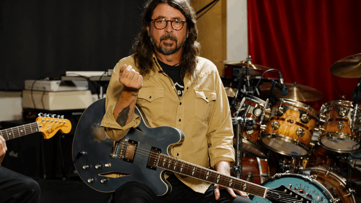 Dave Grohl sits in a studio holding a dark blue semi-hollow electric guitar while gesturing toward the camera.
