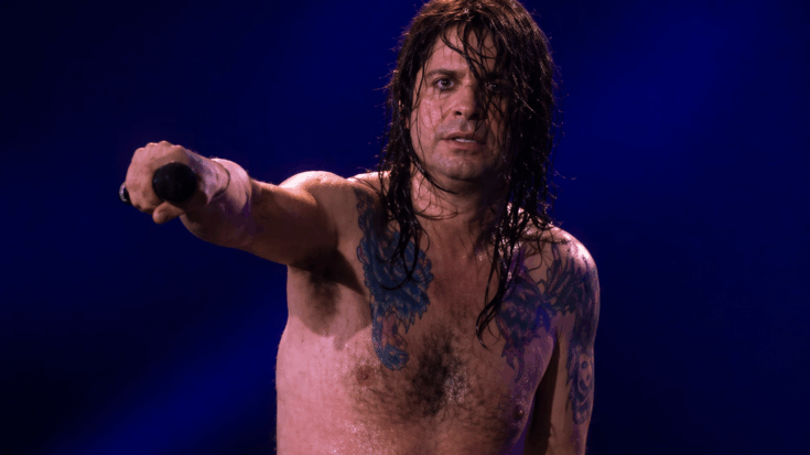 Ozzy Osbourne stands shirtless onstage, dramatically pointing his microphone toward the crowd under deep blue lights.