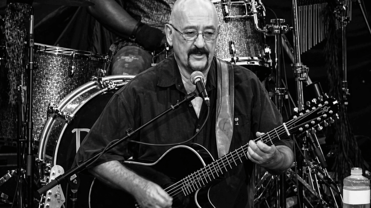 Dave Mason performs at the microphone while holding a black 12-string acoustic-electric guitar.