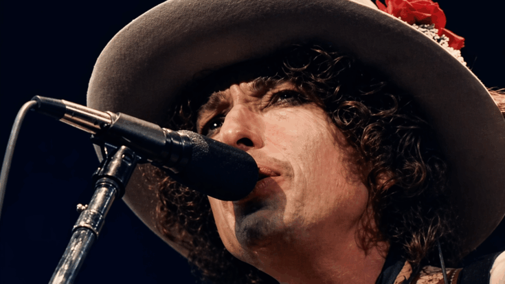 Bob Dylan sings into the microphone in a dramatic close-up, wearing his signature wide-brim hat with a red flower.