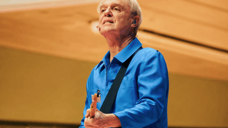 David Byrne performs onstage with his guitar, dressed sharply in a bright blue outfit under warm lights.