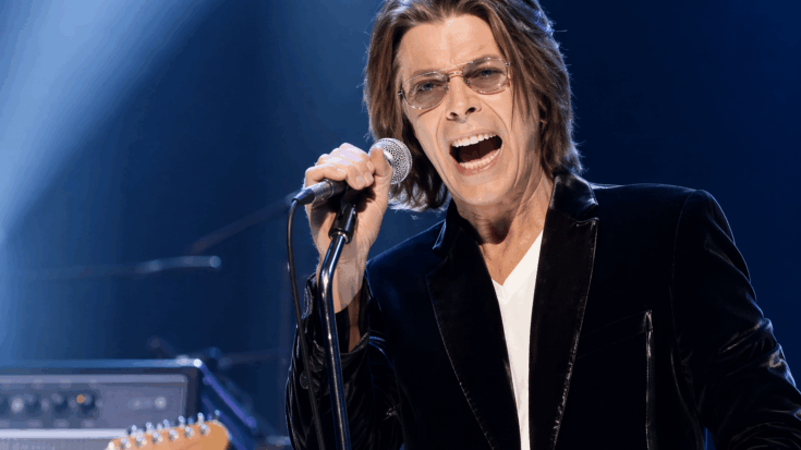 David Bowie sings into a microphone onstage while wearing sunglasses and a dark jacket.