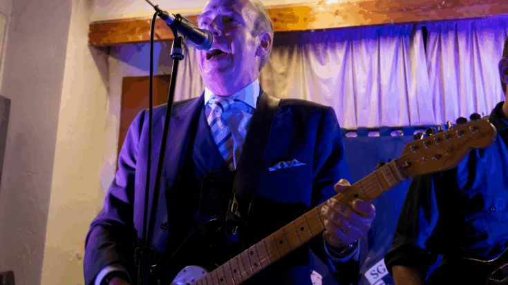 Mick Jones sings into a microphone while playing an electric guitar under blue stage lights.
