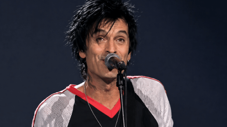 Tommy Lee sings into a microphone onstage while wearing a black and white shirt.