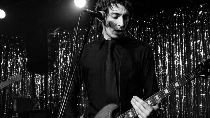 Gregg Foreman plays a red electric guitar onstage beside a microphone under warm lights.