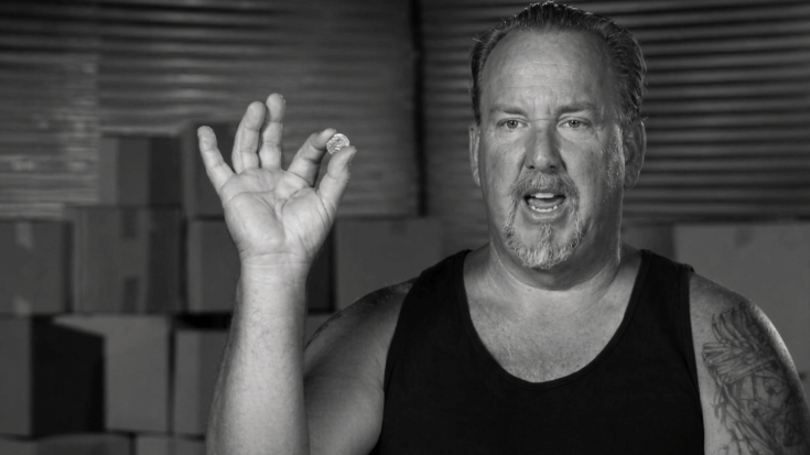 Darrell Sheets holds up a small coin while speaking in front of stacked storage boxes.