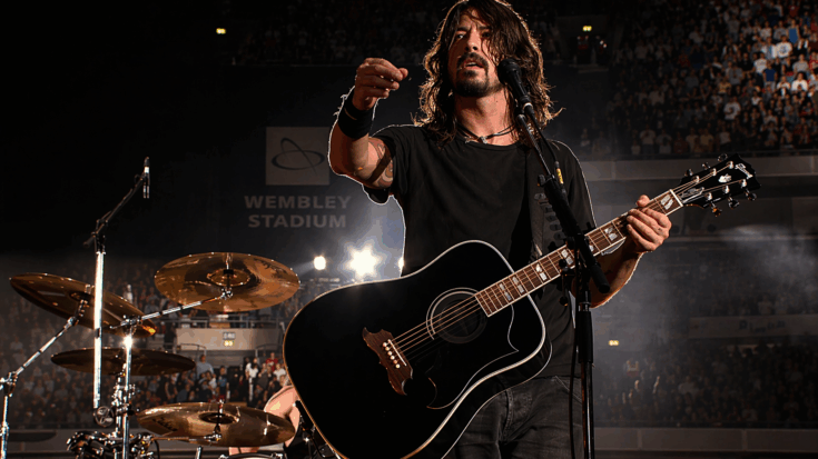 Dave Grohl stands onstage at Wembley Stadium holding a black acoustic-electric guitar.