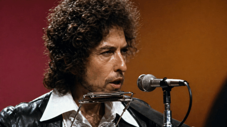 Bob Dylan sings at the microphone with a harmonica holder around his neck in a vintage stage close-up.