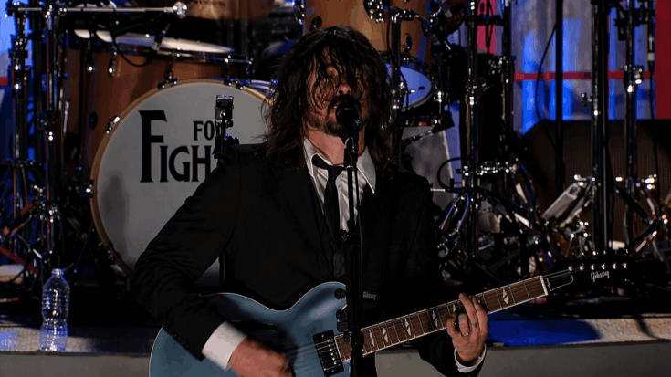 Dave Grohl performs intensely onstage, singing into the mic while playing a blue Gibson electric guitar.