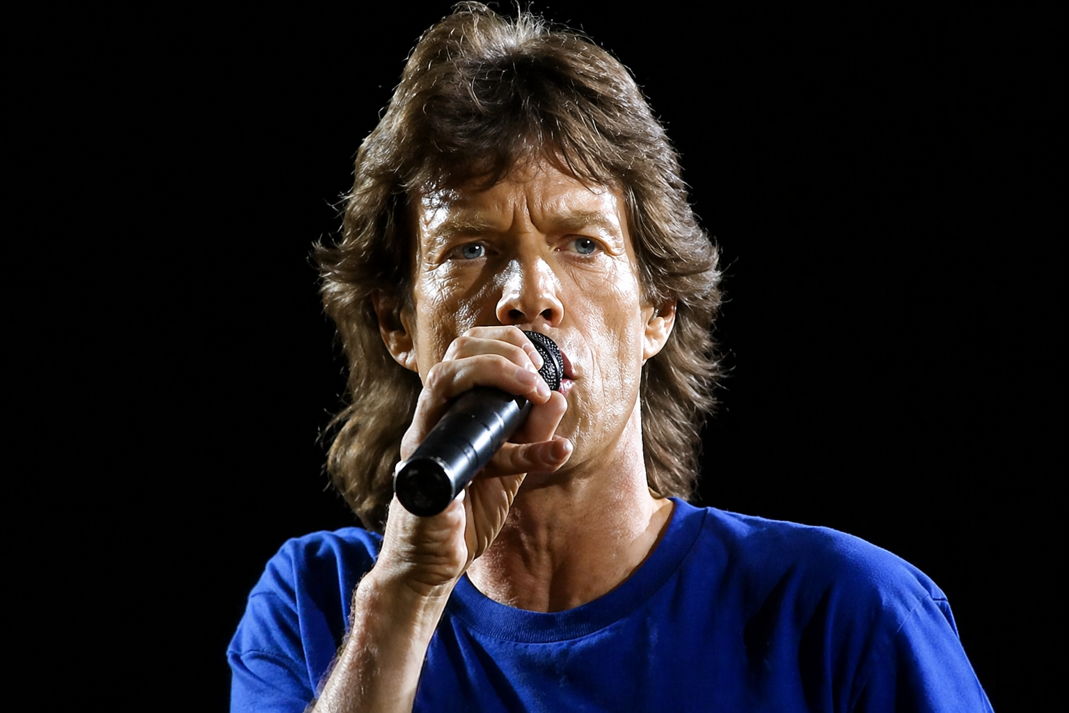 Mick Jagger sings intensely into the microphone in a dramatic close-up against a dark stage backdrop.