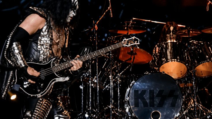 KISS performs onstage as the bassist plays a black electric bass guitar beside the drummer.