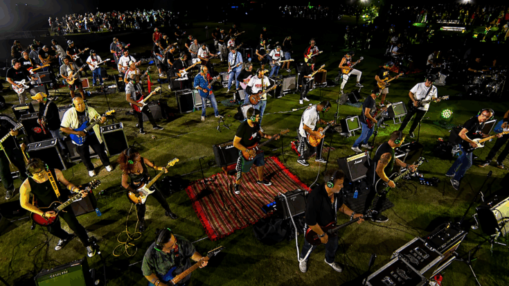 A massive group of musicians perform together outdoors at night, each playing guitars and instruments under bright stage lights.