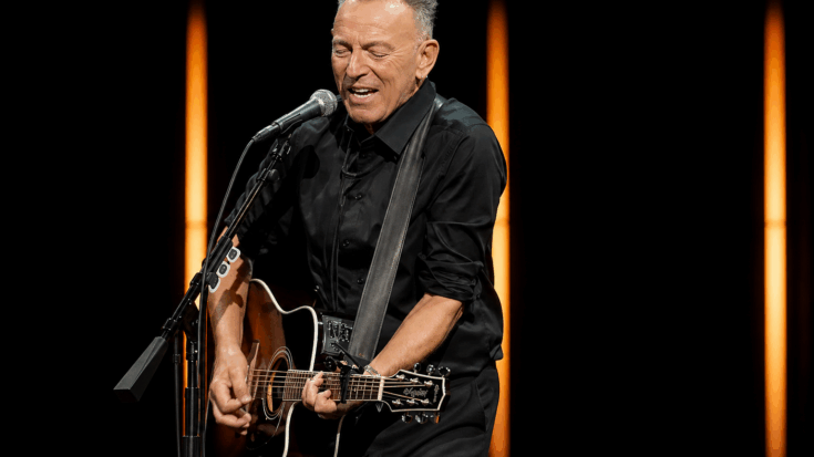 Bruce Springsteen sings into the microphone while playing a black acoustic guitar under warm stage lights.
