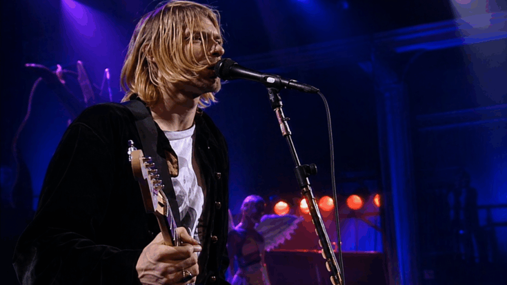 Nirvana performs live as Kurt Cobain sings into the microphone while playing guitar under moody blue stage lights.