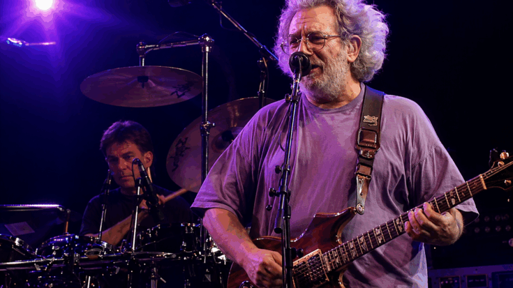 The Grateful Dead perform live under purple stage lights, with the singer playing guitar in front of the drummer.