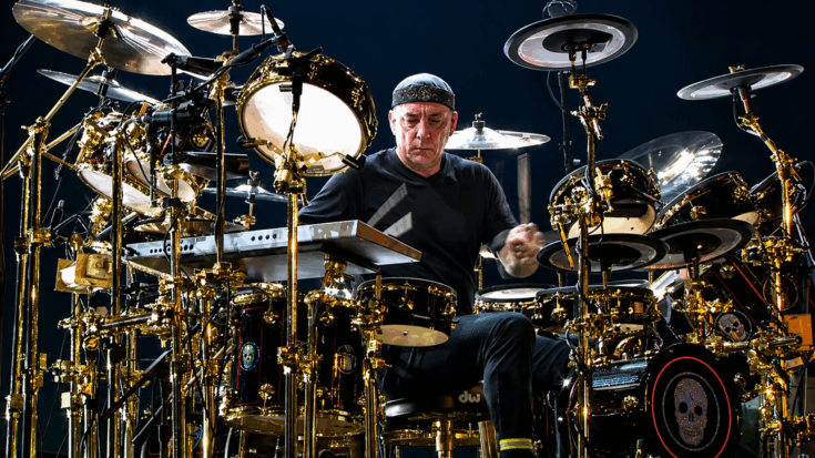 Neil Peart sits intensely behind his massive drum kit, surrounded by gleaming cymbals and percussion gear.