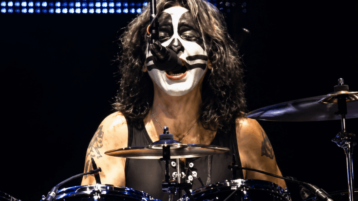 Peter Criss smiles behind the drum kit in full KISS makeup while performing onstage.