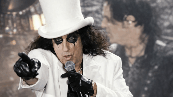 Alice Cooper performs in a striking white outfit and top hat, pointing toward the crowd while singing into the microphone.