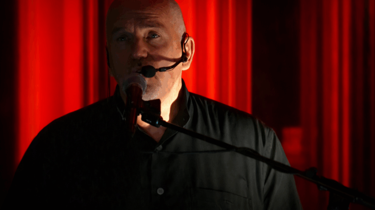 Peter Gabriel sings into a microphone under deep red lighting, creating a dramatic and intimate stage moment.