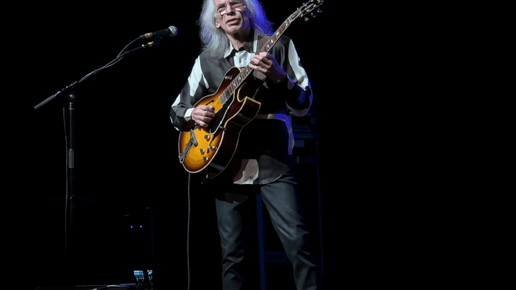 Steve Howe stands under the spotlight, gracefully playing his hollow-body guitar on a dark stage.