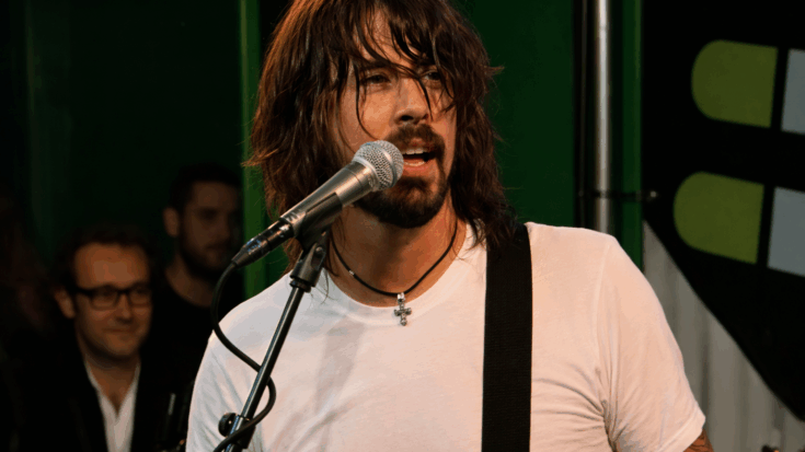 Dave Grohl sings into a microphone with a guitar strap over his shoulder, his shaggy hair partially covering his face under warm stage lights.